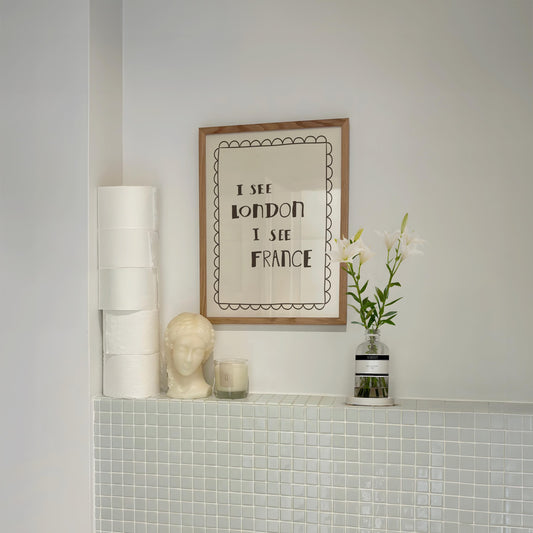 "I See London I See France" funny toilet print displayed on a bathroom shelf with stacked toilet paper, a candle, and a decorative bust