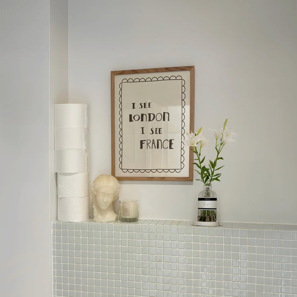 "I See London I See France" funny toilet print displayed on a bathroom shelf with stacked toilet paper, a candle, and a decorative bust
